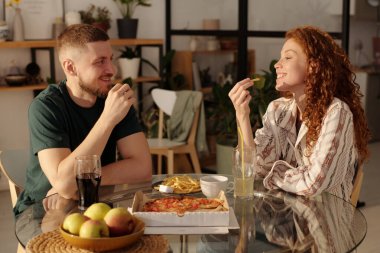 Genç, aç bir çift mutfakta masada oturuyor, patates kızartması yiyor, pizza ısmarlıyor, içki içiyor ve öğle yemeğinin tadını çıkarıyor.