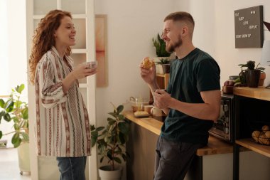 Mutfakta birbirlerinin önünde durmuş sohbet eden ve çay ya da kahveyle donut yiyen mutlu genç çift.