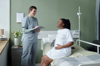 Hemşire hamile Afro-Amerikan bir kadınla nişanlanırken hastane yatağında duruyor. Hastane odasında tıbbi ekipman ve dosya mevcut.