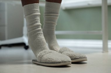 Sıcak terlikler ve rahat çoraplar giyen kişinin yakın planda beyaz duvarla pencerenin yanında dikilirken