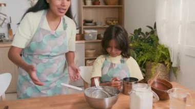 Orta boy İspanyol bir kız mutfak masasında oturup çatal bıçaklara bakarken, annesi birlikte yemek pişirirken bir kase un getirirken.