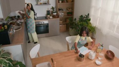 Önlük ve saç bandı takmış İspanyol bir annenin mutfaktaki küçük kızıyla yemek pişirmeye hazırlandığı mutfak dolabından sofraya mutfak malzemesi ve mutfak malzemelerini getirmesi.