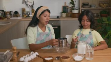 Orta boy İspanyol bir kızın kaşıkları bardağa koyduğu ve annesinin masada oturup modern mutfakta ev yapımı hamur işi yaparken kaşıkları kâseye karıştırdığı bir fotoğraf.