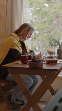 Dikey kenarda, kalın giysiler ve şallar içinde orta yaşlı beyaz bir kadın pencere kenarındaki ahşap masada oturuyor ve Noel tatilinde kır evinde kış manzarası resmi çiziyor.