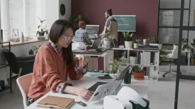 Bilgisayarda, tablette, not yazarken, iş arkadaşlarıyla konuşurken, kahve içerken, bilgisayar başında çalışan, mobil uygulama için kod geliştiren çok ırklı genç bir kadın programcının orta-zaman yan görüntüsü.