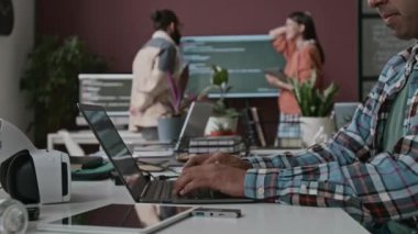 Masada oturan isimsiz bir erkek geliştiricinin orta ölçekli fotoğrafları, dizüstü bilgisayarda bilgisayar kodları yazması, arka planda multimedya ekranında yazılım projesini tartışan ekip üyeleri.