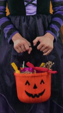 Orta boy dikey eller ve tanınmayan siyah ve mor cadı elbiseli genç bir kızın Cadılar Bayramı şekerleriyle dolu balkabağı kovasıyla stüdyoda turuncu arka planda poz vermesi.