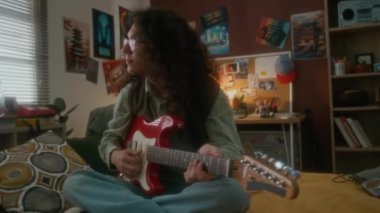 Kabarık saçlı, gözlüklü, kırmızı elektro gitar çalan genç gitarist dolu bir fotoğraf. Dekorasyonlu retro-stil bir odada, yatakta oturuyor, nostaljik 90 'larda.