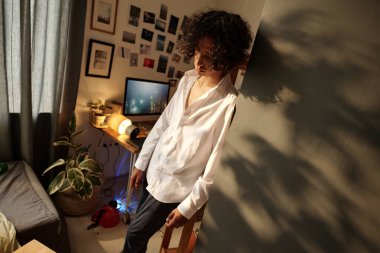 Beyaz tişörtlü sarhoş genç adam baş ağrısı, sarhoşluk ve mide bulantısı çekerken ev ortamında köşede dikiliyor.