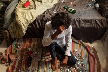 Baş ağrısı ve bulantısı olan genç sarhoş bir adam içki içtikten sonra dağınık yatak odasının zemininde çizgili bir halının üzerinde oturuyor.