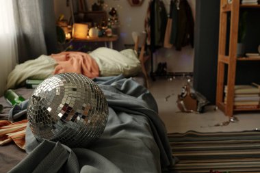 Parlayan disko topu kırışık yatakta yatıyor gri battaniye, giysi, boş şişeler ve diğer şeyler dağınık odanın iç kısımlarına karşı.