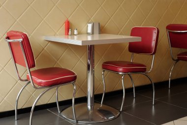Tuz, biber ve ketçapla aksanlı küçük bir masanın etrafında şık kırmızı sandalyelerin olduğu klasik restoran sahnesi. Elmas desenli duvar retro titreşimi tamamlıyor