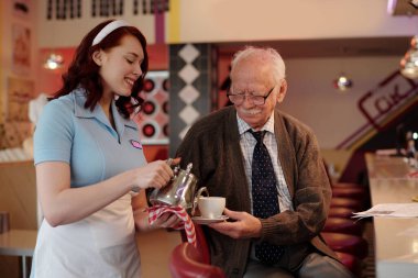 Gülümseyen garson retro lokantada oturan yaşlı adama kahve servisi yapıyor. Genç garson ile yaşlı müşteri arasındaki etkileşim sıcak ve nostaljik bir atmosfer yaratıyor.