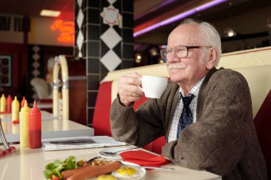 Elderly man savoring cup of coffee while seated in retro diner, showcasing a nostalgic ambiance with classic decor, surrounded by breakfast items and reading material clipart