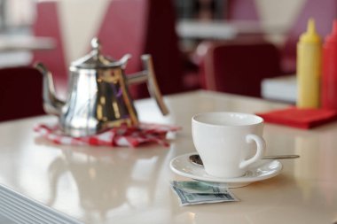 Parlak çaydanlık ve beyaz bardak, arka planda baharatlar, restoran ortamında sıradan yemek atmosferini yansıtıyor.