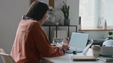Orta boy, turuncu kazaklı genç Asyalı kadın programcının masada oturup dizüstü bilgisayara yazılım kodları yazarken ve ofiste tek başına çalışırken tabletten kontrol ederken görüntüsü.
