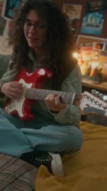 Dikey çekimde kabarık saçlı, gözlüklü, eski tarz yatak odasında yatağında oturan, kırmızı elektro gitar çalan, tasasız ve nostaljik 90 'lı yılların beyaz çocuğu.