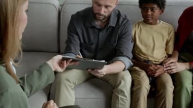 Kadın sosyal hizmet görevlisinin, ev teftişi sırasında beyaz karı-kocaya evlatlık belgeleri verirken mutlu çiftin evrakları imzalarken ve Afrikalı Amerikalı üvey oğluna sarılırken orta boy fotoğrafı.