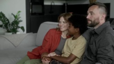Orta ölçekli mutlu beyaz koruyucu aile, baba, anne ve Afrikalı Amerikalı evlat edinen çocuğun el ele tutuşup TV 'de komik çizgi filmler izlediği fotoğraf.