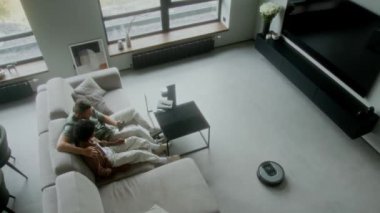 Geniş açılı robot süpürgesi oturma odasında yerleri süpürüyor, Afrikalı-Amerikalı evlatlık oğlu olan beyaz bir koruyucu baba koltukta dinleniyor, akıllı telefon ile akıllı alet kullanmayı öğreten bir adam.