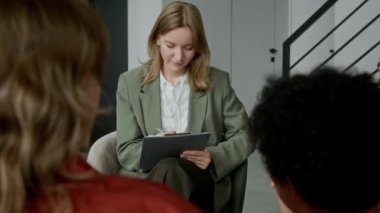 Orta ölçekli gülümseyen beyaz kadın sosyal hizmet görevlisi koruyucu aile ziyareti sırasında koruyucu ailenin evlat edinen siyahi ebeveynleriyle konuşurken, sağlıkla ilgili sorular sorarken ve rapora cevaplar yazarken.