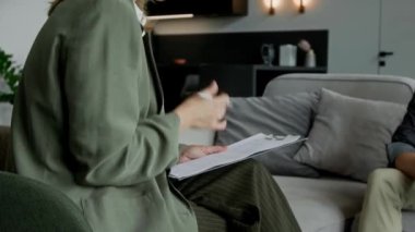 Genç beyaz kadın sosyal hizmet görevlisinin evdeki koltuğunda otururken, kontrol sırasında aile üyeleriyle röportaj yaparken, rapor için panoya kontrol listesi doldururken orta eğik bir yan görüntüsü.