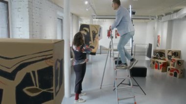 Beyaz kadın sanatçı ve galeri müdürünün, yaklaşan çağdaş sanat sergisi için hazırlanırken, holdeki tavana konseptsel sanat eserlerini astığı tam zoom çekimi.