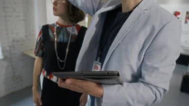 Beyaz tenli erkek sergi küratörünün dijital tablet üzerinde sanat eserleri için şık kadın sanatçı düzenleme planı, kadının başını sallaması ve gülümsemesi, kurulum hazırlarken orta boy yakın çekimi