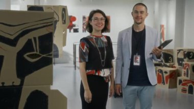Orta boy beyaz çağdaş kadın sanatçı, şık renkli kıyafetler içinde ve gri takım elbiseli erkek galeri müdürü, modern sanat sergisinde ayakta duran, gülümseyen, kameraya bakan