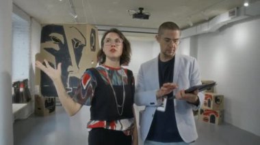 Sanat eserlerinin düzenlenmesi için kişisel sergi hazırlarken galeri müdürüyle konuşan, sanatsal görüşü açıklayan, tablet üzerine notlar alan beyaz kadın sanatçı.