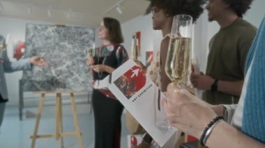Çok ırklı bir grup ziyaretçi, ellerinde şampanya flütleri, sehpanın etrafında soyut tabloyla dikilip sanat eserlerini sunan kadın sanatçıları dinliyorlar.