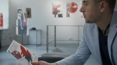 Orta ölçekli, kırpık saçlı, elinde broşür olan beyaz bir adamın galeri ya da müzedeki çağdaş sergide sergilenen sanat eserlerini okuyup izlediği bir fotoğraf.