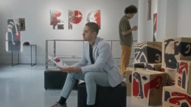 Müzede ya da galeride sergilenen sanat eserlerini izleyen iki farklı erkek ziyaretçi, puffe üzerinde broşürle oturan beyaz bir adam ve akıllı telefondan bilgi arayan Orta Doğulu bir adam.