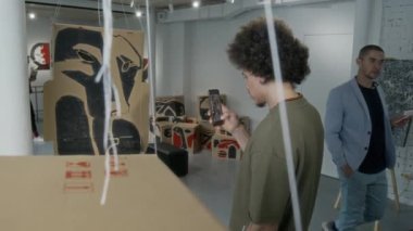 Orta boy, kıvırcık saçlı, modern sanat galerisinde boyalı karton kutuların kavramsal kurulumunun önünde duran ve akıllı telefonuyla fotoğraf çeken Orta Doğulu genç bir adam.