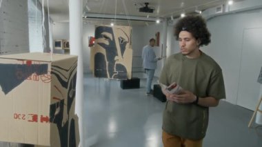 Orta boy Orta Doğulu genç bir adam müzede çağdaş sanat sergisi etrafında dolanıyor, kavramsal karton tesisatına uğruyor, üzerinde çalışıyor ve sakalını okşuyor.