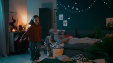 Küçük yaramaz beyaz kız Noel ağacına doğru koşuyor ve gizlice hediyesini açıyor. Büyükbaba oturma odasında kanepede uyurken, tatil beklentisiyle.
