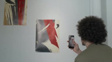 Modern sanat sergisinde beyaz duvarda sergilenen kalın, kısa kıvırcık saçlı ve akıllı telefonuyla fotoğraf çeken genç bir adamın orta boy arka çekimi.