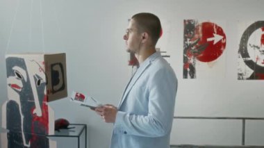 Gri takım elbiseli, elinde broşür, sergideki sanat eserlerine doğru yürürken, kulaklıkları ses rehberiyle ayarlarken orta ölçekli beyaz erkek ziyaretçi fotoğrafı.