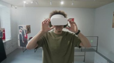 Orta boy kıvırcık saçlı, sanal gerçeklik gözlükleri takan genç bir Orta Doğulu adamın sanat galerisindeki sergilere katılırken görüntüsü.