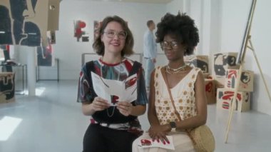 Modern sanat müzesindeki bankta oturan, kameraya bakıp gülümseyen ve sergiden keyif alan iki zarif beyaz ve Afrikalı Amerikalı kadının orta boy portresi.