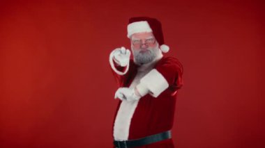 Geleneksel kostümlü, kendine güvenen Noel Baba 'nın kırmızı arka planda garip ve modern dans figürleri yaparken ve kameraya, şablona ve uzay kopyalarına bakarken orta ölçekli bir fotoğraf.