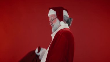 Geleneksel kostümlü arsız ve kendine güvenen Noel Baba 'nın yavaş yavaş portresi omuzlarına bir çuval hediye koyar ve kırmızı arka planda kameraya bakar. Şablon için uygun, alanı kopyala