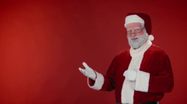 Geleneksel kostümlü kendine güvenen Noel Baba 'nın orta boy portresi kırmızı arka plandaki bağlantıyı ya da ürünü işaret ediyor ve başparmaklarını kaldırıyor, kameraya bakıyor ve gülümsüyor. Şablon, boşluğu kopyala