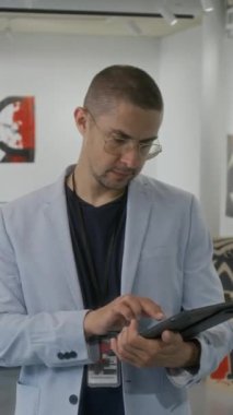 Gri takım elbiseli, rozetli, dijital tablette yazan, modern sanat sergisinde çalışan, kameraya bakan beyaz erkek müze müdürünün orta boy dikey portresi.