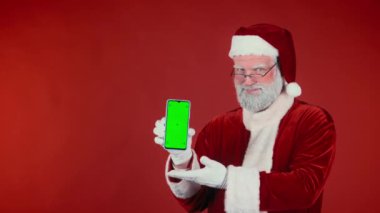 Modern Noel Baba 'nın geleneksel kostümlü orta boy portresi akıllı telefonu yeşil renkli şablon ekranla göstererek, ürünü önererek, kırmızı arka planda başparmaklarını göstererek, uzayı kopyalayarak,