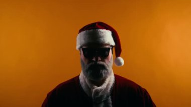 Güneş gözlüklü, geleneksel kostümlü, gümüş zincirli klasik rockçı Noel Baba 'nın göğüs portresi kameraya bakıyor, boynuz el işareti yapıyor ve sarı arka planda neşeli müziğe doğru ilerliyor.