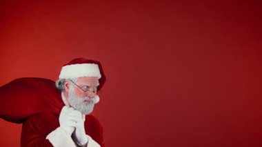 Geleneksel kostümlü sessiz Noel Baba 'yı göğsünden bıçaklayıp Noel zamanı kırmızı arka planda uslu çocuklara dağıtmak için bir çuval hediyeyle geçerken çek. Şablon, boşluğu kopyala