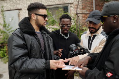 Açık şehir ortamındaki belgeleri incelerken sohbet eden bir grup genç adam, bazı kameralar ve cihazlar, gelişigüzel giyinmiş, dostluk ve takım çalışmasını gösteriyor.