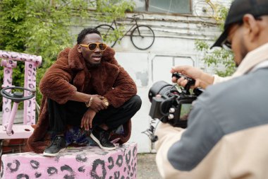 Kel adam kahverengi kürklü poz verirken fotoğrafçı arka bahçedeki profesyonel kamera ekipmanlarını kullanarak çarpıcı görsel etki yaratıyor.