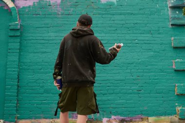 Siyah kapüşonlu genç adam, boya sıkmadan ve sanat eseri yaratmadan önce yeşil tuğla duvarın önünde dikiliyordu.